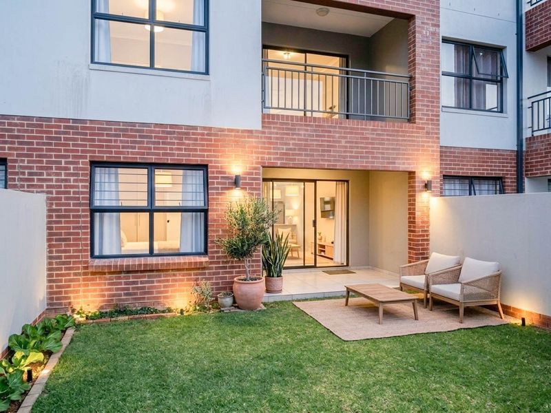 A well-maintained rear exterior view of an apartment featuring a private garden, tiled patio area, and a balcony with modern brickwork.