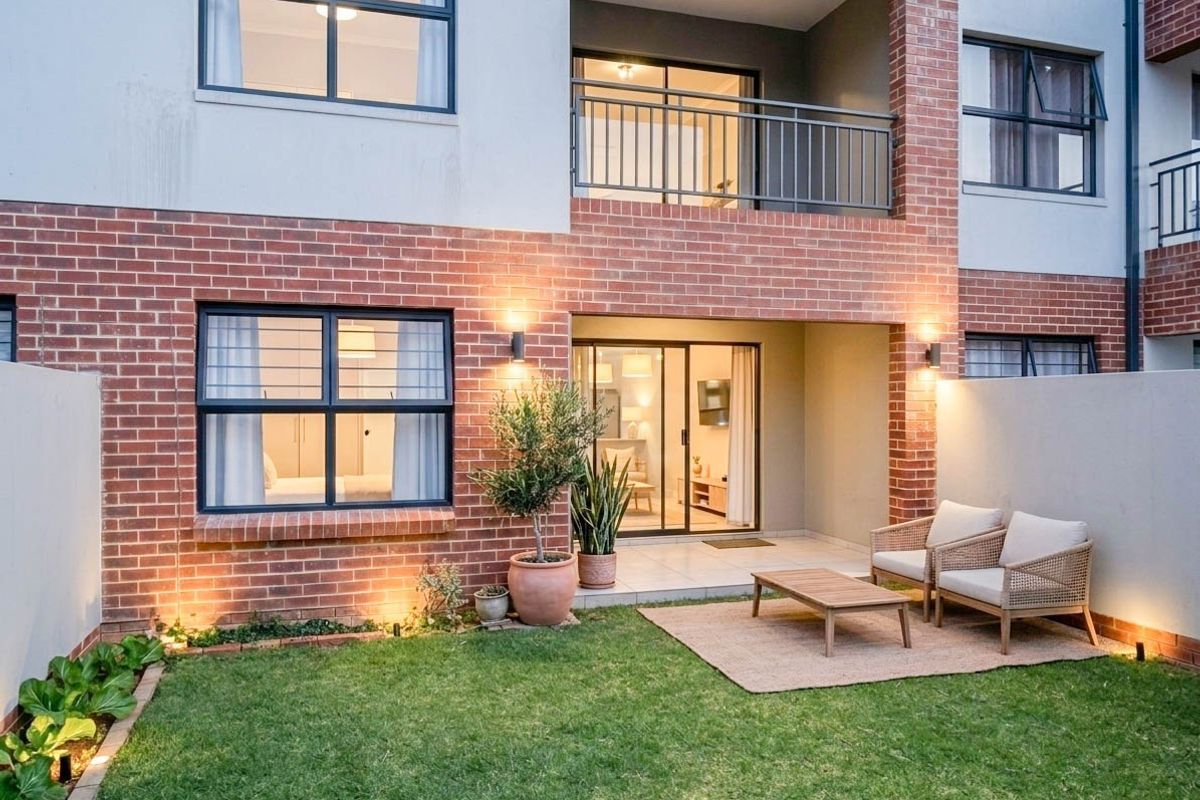 A well-maintained rear exterior view of an apartment featuring a private garden, tiled patio area, and a balcony with modern brickwork.