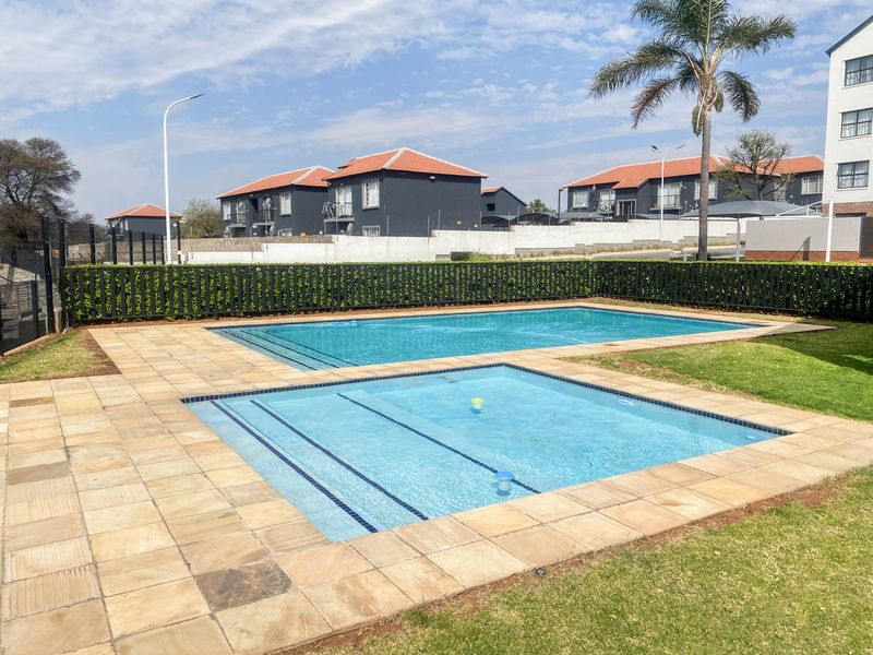 A well-maintained communal swimming pool area featuring paved surrounds, manicured lawn, and secure perimeter fencing within a residential apartment complex.