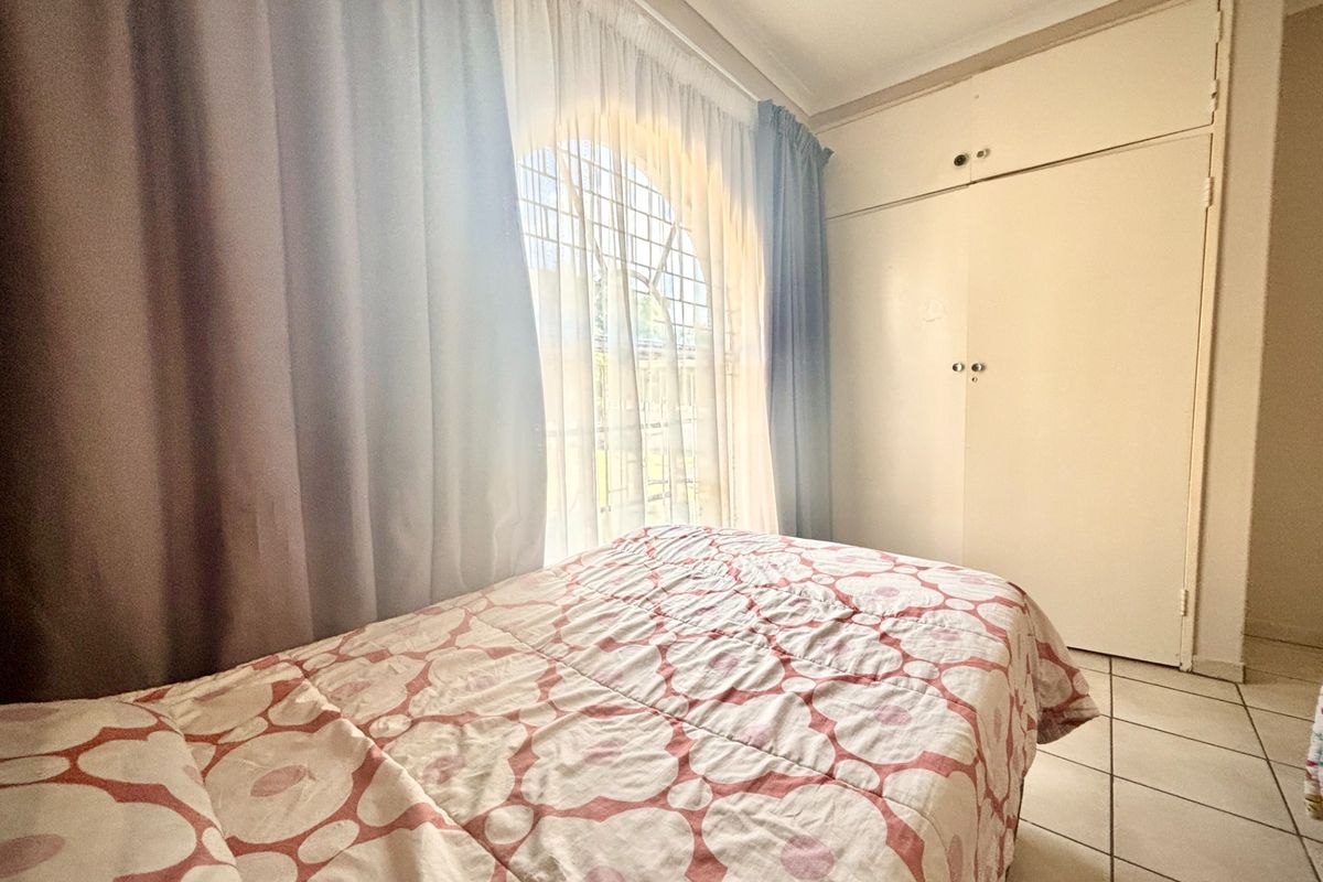 A bedroom featuring built-in cupboards, tiled flooring, and an arched window with security bars, offering a functional and well-lit living space.
