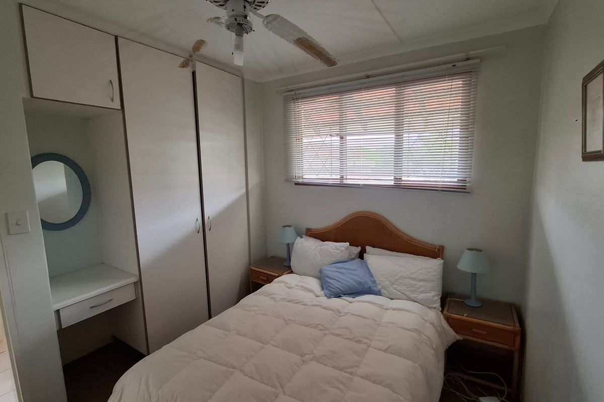 A functional bedroom featuring built-in cupboards, a vanity area, and a ceiling fan, with natural light provided by a window with venetian blinds.
