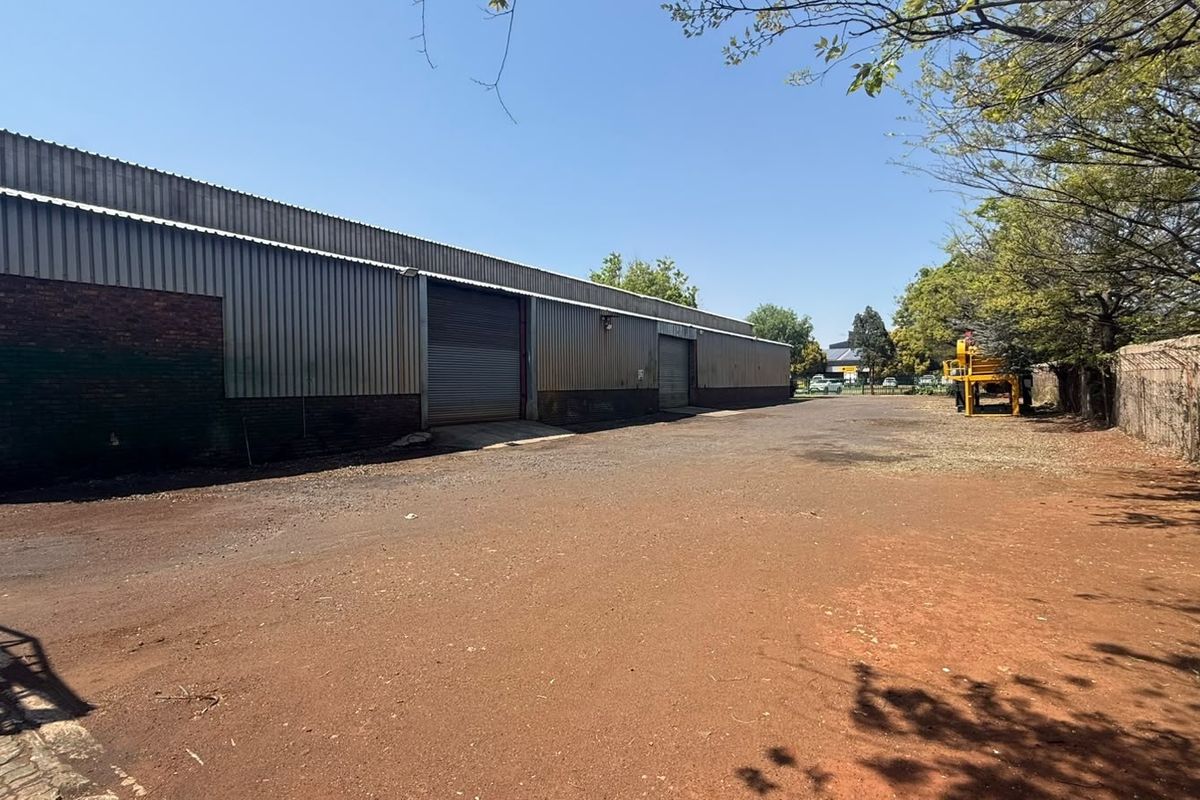 Rear exterior view of an industrial warehouse featuring corrugated metal cladding, roller shutter doors, and a large unpaved yard area.
