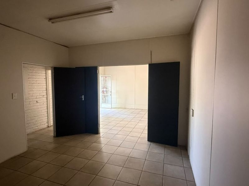 An empty, functional office space featuring tiled flooring, fluorescent lighting, and double doors leading to an adjacent room with exposed brick detailing.