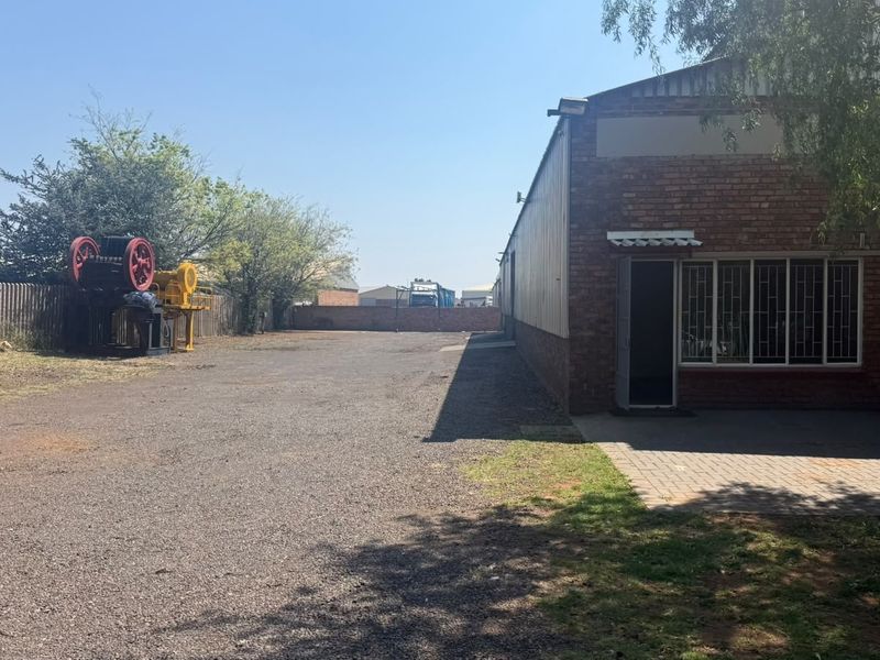 Rear exterior view of the industrial property featuring a brick facade, security-barred windows, a paved patio section, and a large gravel yard area.