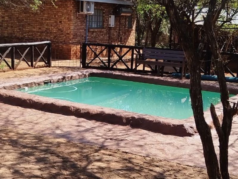 Outdoor pool area featuring a paved surround, brick building facade, and timber fencing, set within a natural garden environment.