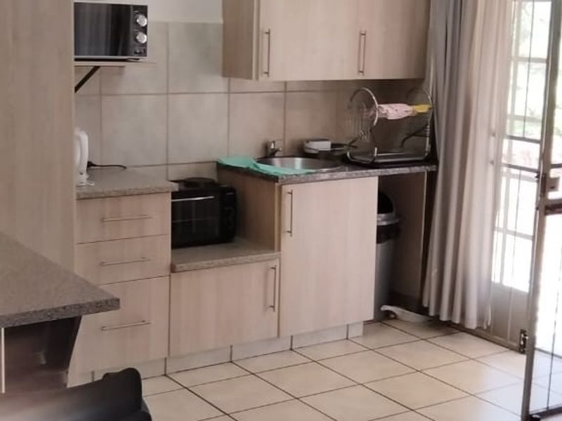 A functional kitchen featuring light wood-finish cabinetry, laminate countertops, a stainless steel sink, and tiled flooring with direct access to an outdoor area.