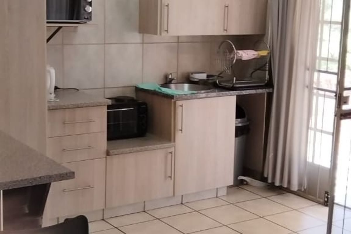 A functional kitchen featuring light wood-finish cabinetry, laminate countertops, a stainless steel sink, and tiled flooring with direct access to an outdoor area.