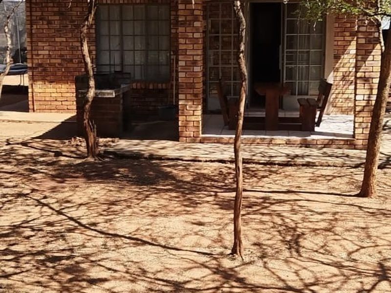 Rear view of a brick house featuring a covered patio area and a built-in braai, set within a natural garden environment.