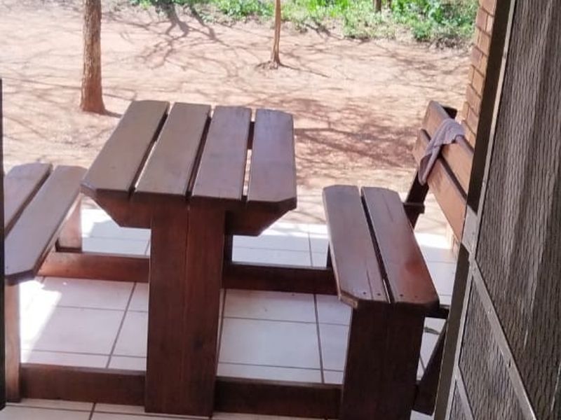 A covered outdoor patio area featuring tiled flooring and exposed brickwork, overlooking a natural bushveld garden setting.