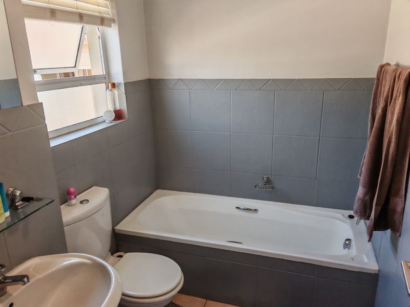 A functional bathroom featuring a built-in bathtub, pedestal basin, toilet, and neutral grey wall tiling with a window for natural light.