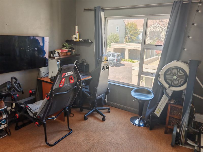A bedroom currently utilized as a gaming and fitness room, featuring carpeted flooring and a large window with views of the complex.