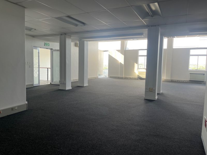 A spacious, open-plan office area featuring neutral carpeting, structural columns, suspended ceilings with recessed lighting, and large windows providing natural light.