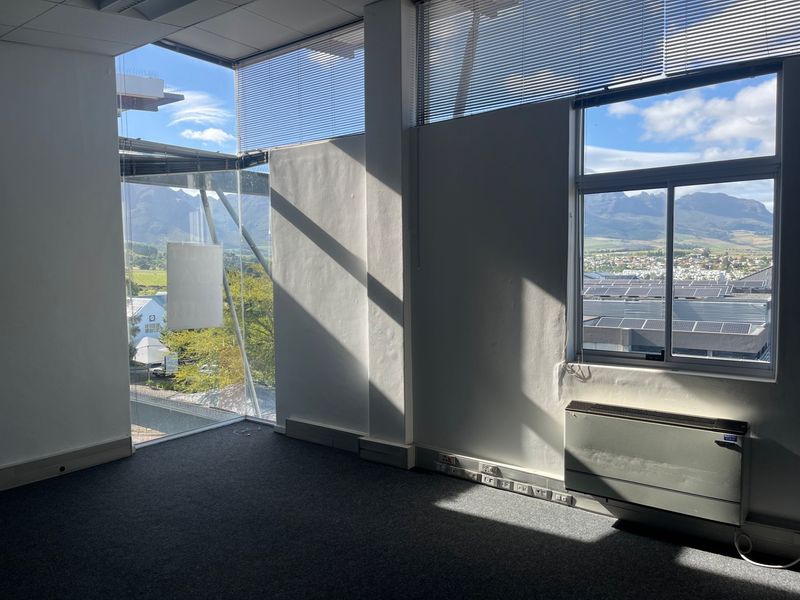 A bright, empty office space featuring large windows with mountain views, carpeted flooring, integrated HVAC unit, and functional power skirting.