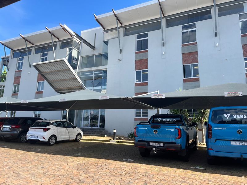 Front elevation of a modern multi-story office building featuring a paved parking area with shade cloth carports and glass entrance.