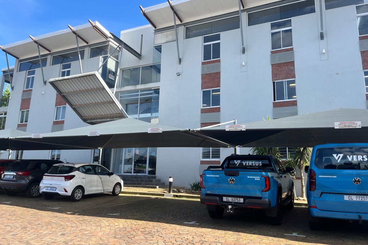 Front elevation of a modern multi-story office building featuring a paved parking area with shade cloth carports and glass entrance.