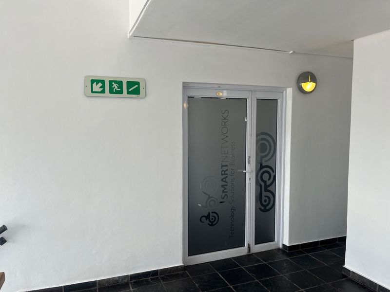 A clean office entrance featuring a glass door with frosted branding, tiled flooring, and a wall-mounted emergency exit sign.