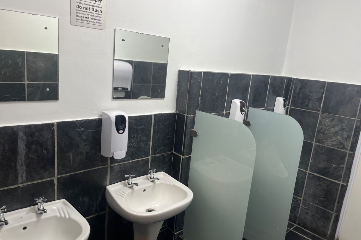 A functional office bathroom featuring pedestal wash basins, frosted glass privacy screens, and dark tiled wall and floor finishes.