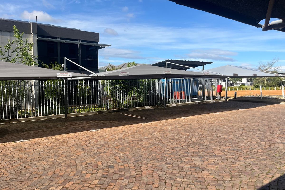 A paved outdoor parking area featuring shade sail structures and secure metal fencing, adjacent to the office building exterior.