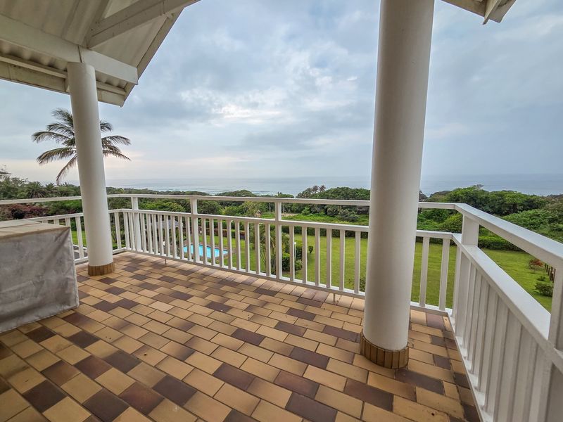 A spacious covered balcony featuring tiled flooring, white railings, and support columns, offering scenic views of the surrounding garden and ocean.