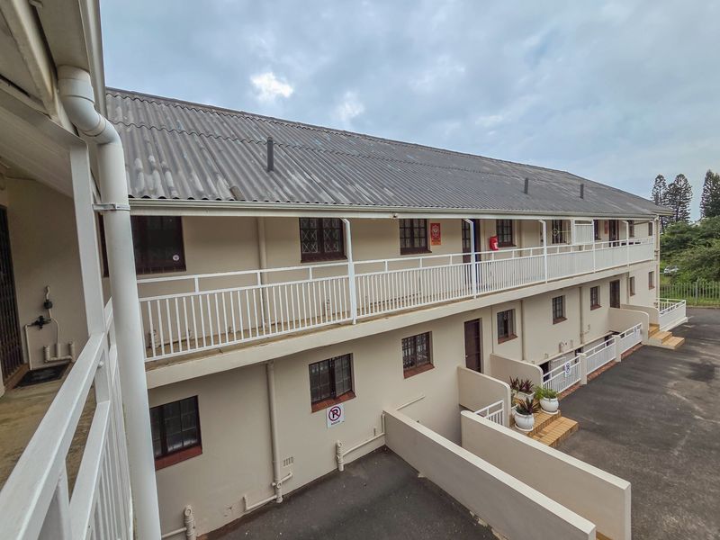 Exterior view of a multi-story apartment building featuring metal balustrades, a corrugated roof, and a paved driveway area.