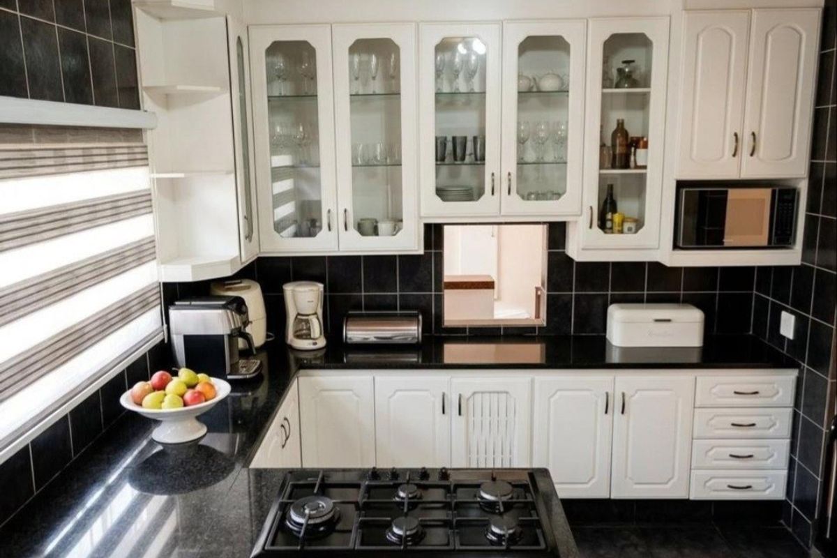 A well-maintained kitchen featuring white cabinetry, black granite countertops, a gas hob, and a striking black tiled backsplash with timber ceiling accents.