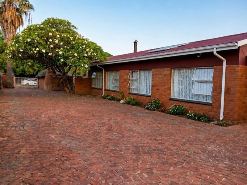A well-maintained brick house exterior featuring a paved driveway, metal roofing with solar panels, and established garden landscaping.