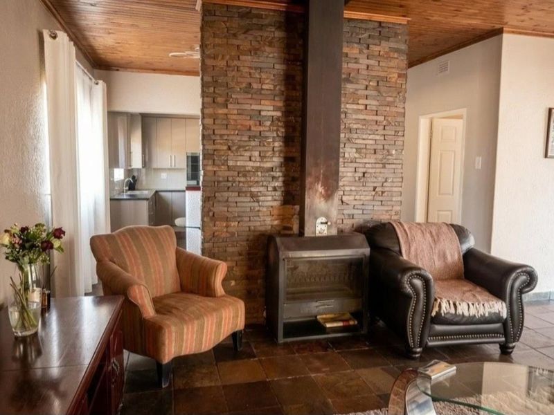 Open-plan living area featuring a prominent stone fireplace with a wood-burning stove, timber ceilings, and tiled flooring, with a view into the kitchen.