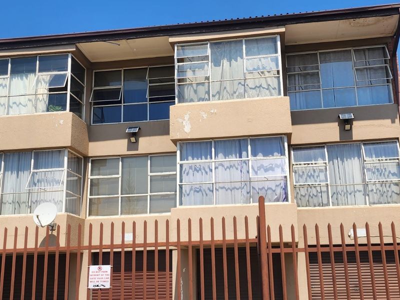Front elevation of a multi-story apartment building featuring metal security fencing, roller shutter garage doors, and aluminium window frames.