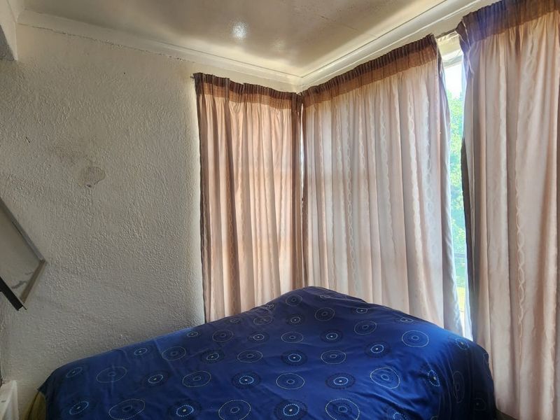 A bedroom featuring a corner window with natural light and textured wall finishes requiring maintenance.
