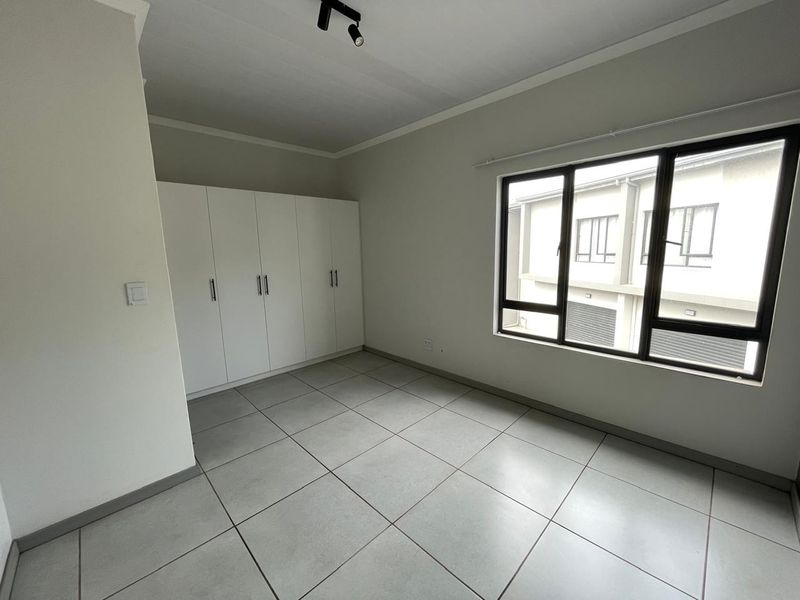 A bright, empty bedroom featuring neutral tiled flooring, built-in white cupboards, and large aluminium-framed windows providing natural light.