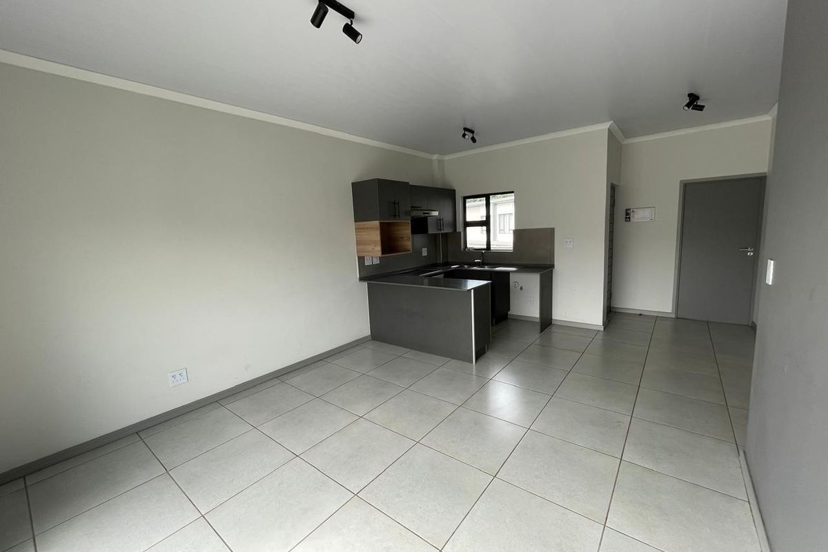 An open-plan living and kitchen area featuring modern grey cabinetry, tiled flooring, and contemporary track lighting in a clean, vacant space.