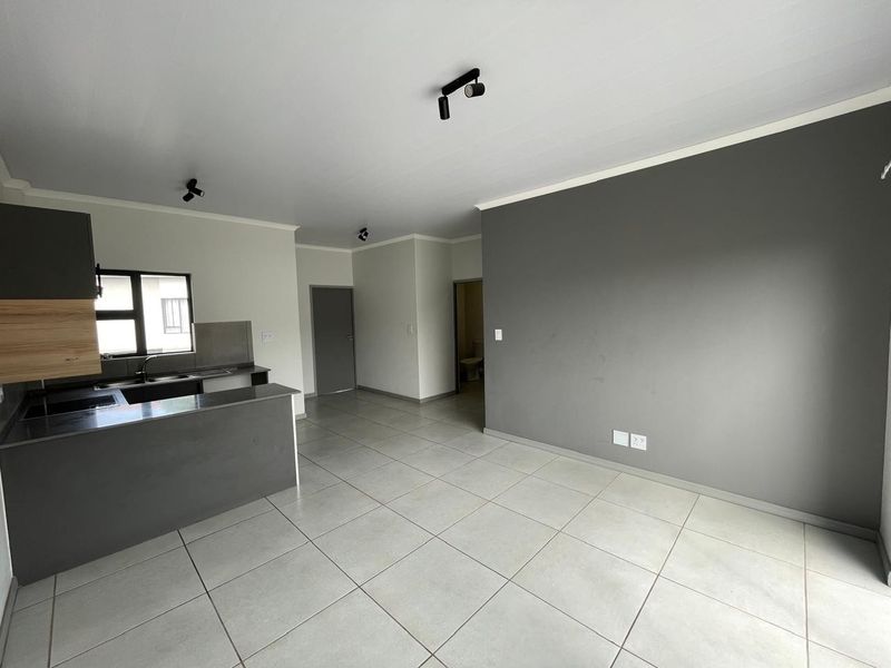 An open-plan living and kitchen area featuring modern grey cabinetry, neutral tiled flooring, and contemporary track lighting in a clean, vacant space.