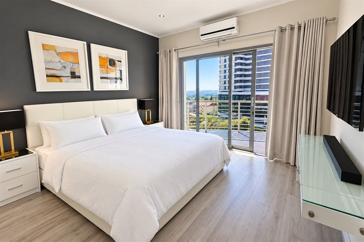 A modern, well-maintained bedroom featuring laminate flooring, an air conditioning unit, and a sliding glass door leading to a private balcony with city views.