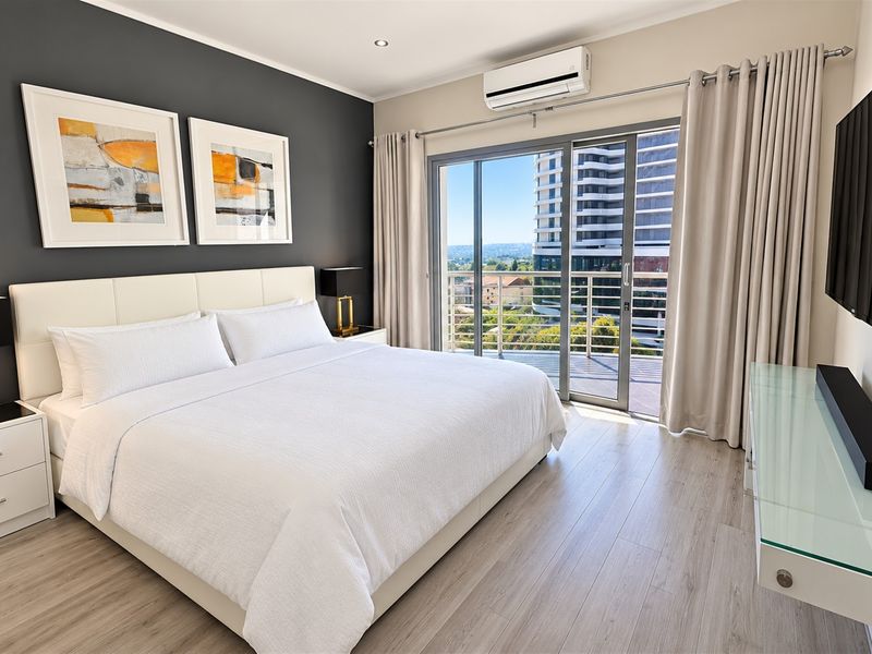 A modern, well-maintained bedroom featuring laminate flooring, an air conditioning unit, and a sliding glass door leading to a private balcony with city views.