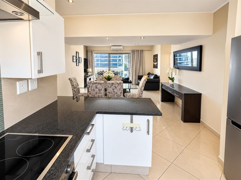 Modern open-plan kitchen and living area featuring granite countertops, sleek cabinetry, tiled flooring, and seamless flow to a balcony through sliding glass doors.