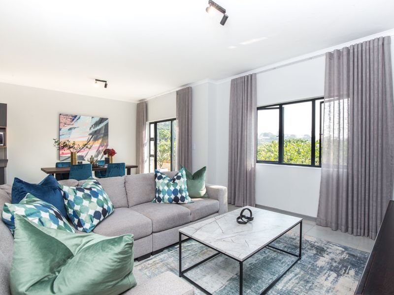 A bright open-plan living and dining area featuring tiled floors, modern track lighting, and large windows with views of the surrounding greenery.