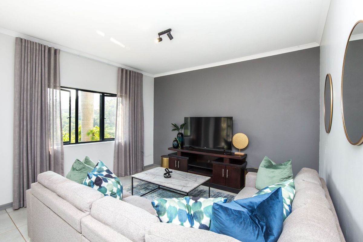 A well-maintained living room featuring a dark grey accent wall, neutral tiled flooring, and large windows with modern curtain tracks.