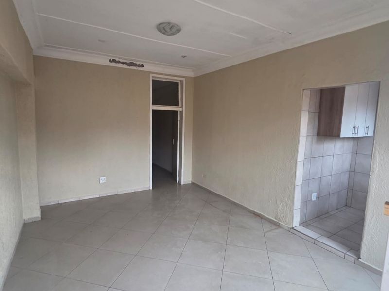 An empty living room featuring tiled floors and a connected kitchen area, requiring minor cosmetic repairs to the wall plaster and lighting.