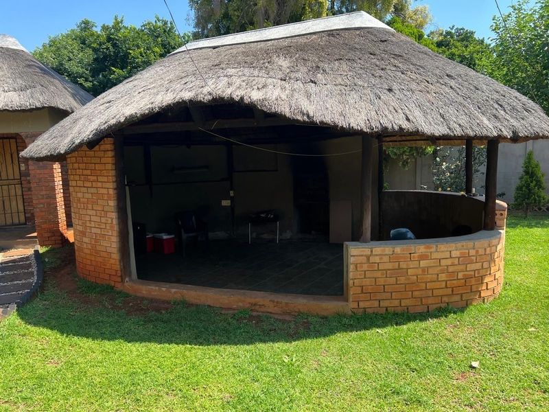 An outdoor thatched lapa area featuring a built-in braai, brick masonry walls, and tiled flooring, set within a garden environment.