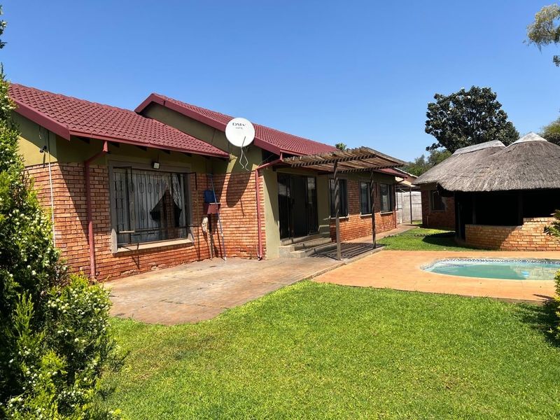 Rear view of the property featuring a brick facade, tiled roof, wooden pergola, swimming pool, and a traditional thatched lapa in the garden.