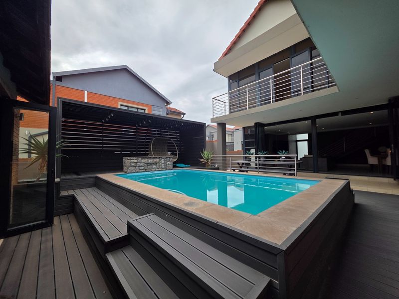 A modern outdoor entertainment area featuring a raised swimming pool with composite decking, a stone-clad water feature, and a stylish black timber pergola.