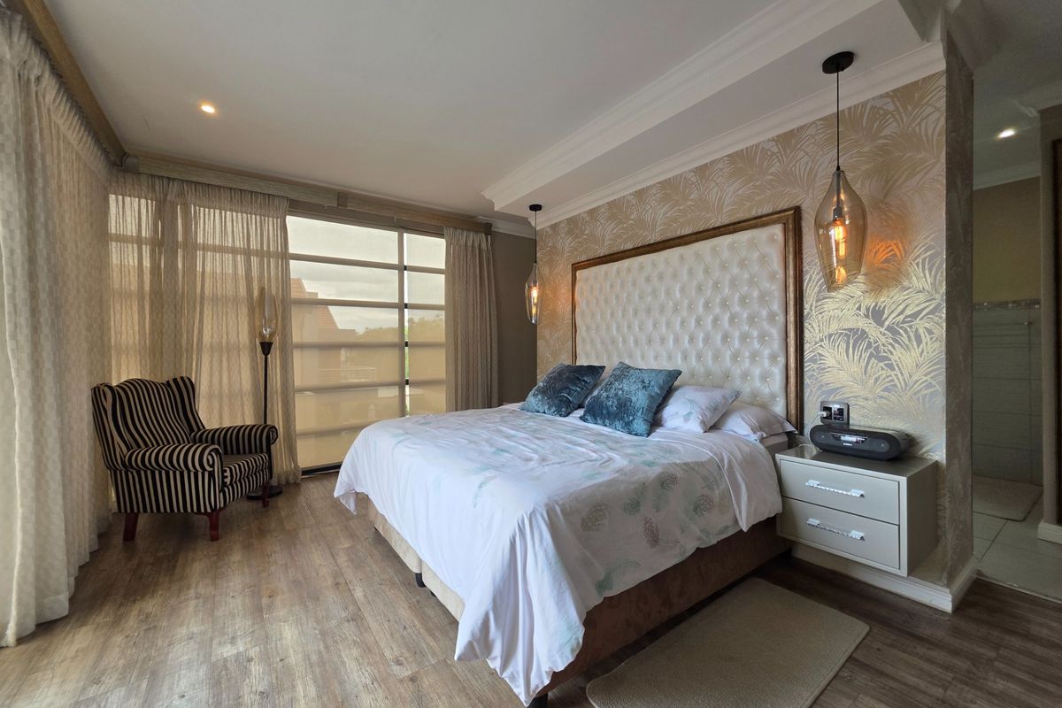 A well-appointed bedroom featuring laminate flooring, decorative wallpaper, recessed ceiling details, and modern pendant lighting with access to a private balcony.