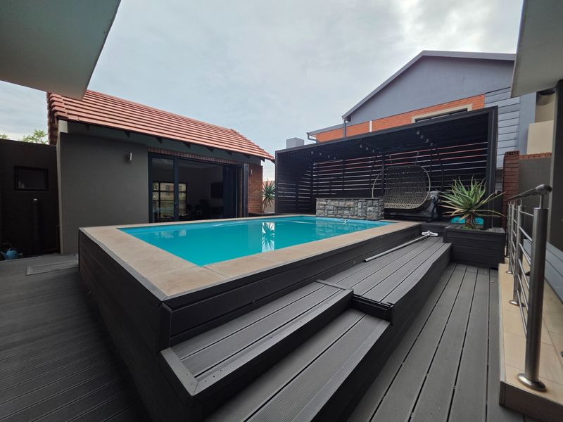 Modern outdoor entertainment area featuring a raised swimming pool, composite decking, and a stylish timber pergola with a stone water feature.