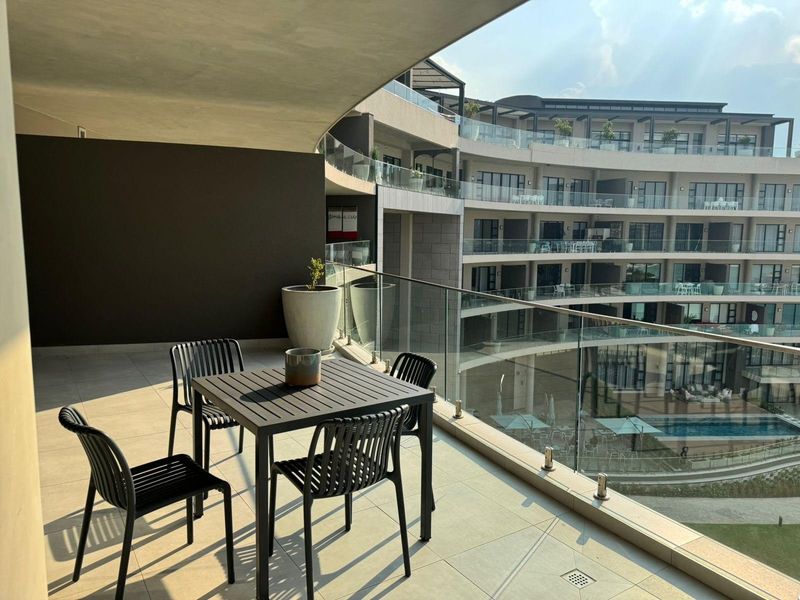 Spacious covered balcony featuring a modern glass balustrade and tiled flooring, offering views of the complex architecture and pool area.