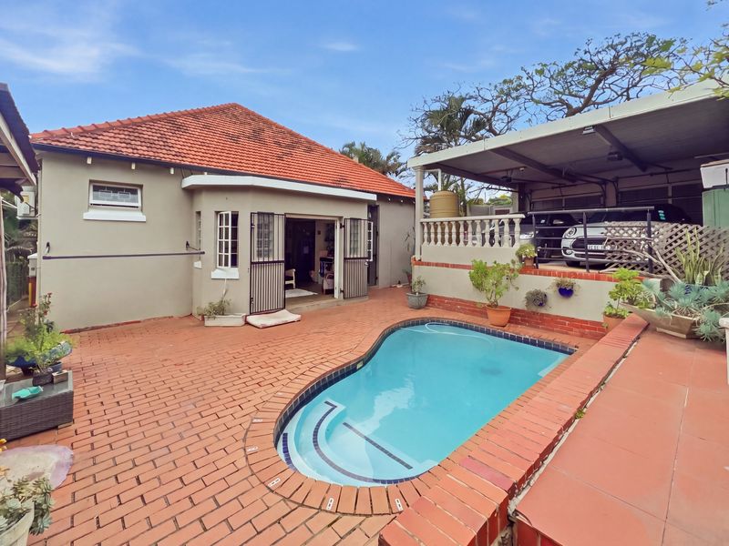 Rear exterior view featuring a swimming pool, brick-paved patio area, and an attached carport with a balustrade.