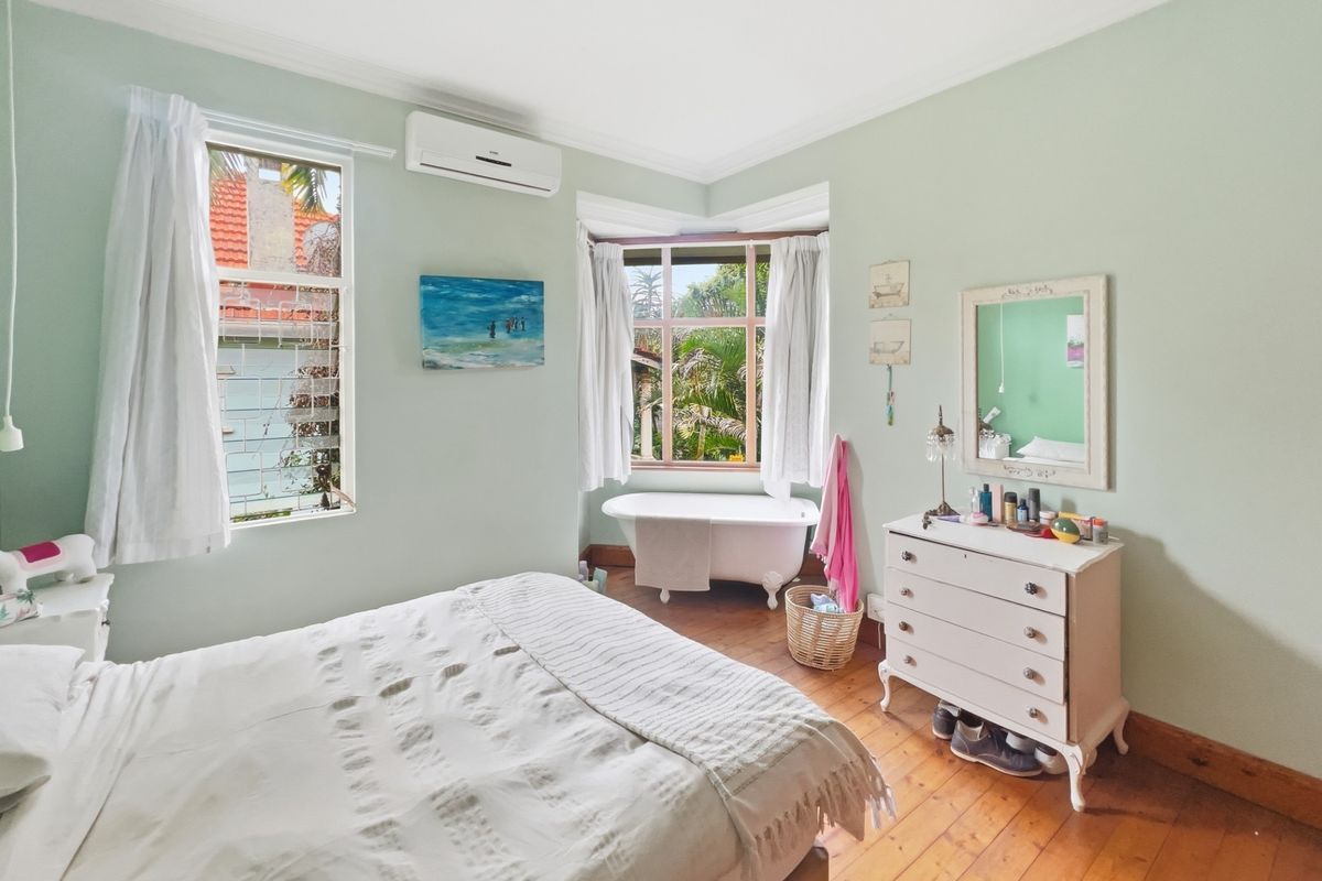 A spacious bedroom featuring polished timber flooring, a classic clawfoot bathtub positioned in the bay window, and a wall-mounted air conditioning unit.