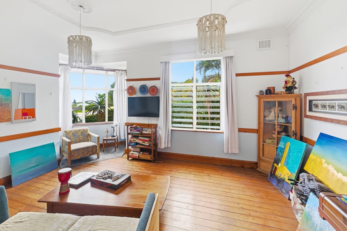 A bright living room featuring original timber flooring, decorative ceiling roses, picture rails, and large windows providing natural light and garden views.
