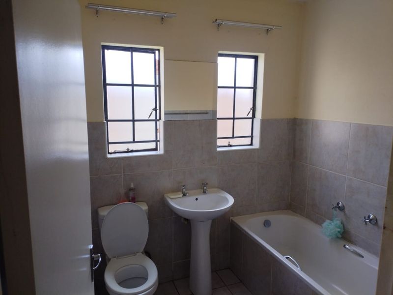 A functional bathroom featuring a pedestal basin, toilet, and bathtub with neutral wall tiling and natural light from two steel-framed windows.