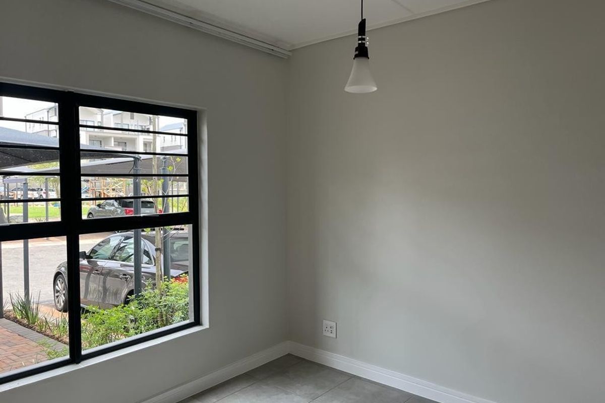 A bright, empty living area featuring modern tiled flooring, neutral walls, and large black aluminium windows providing natural light.