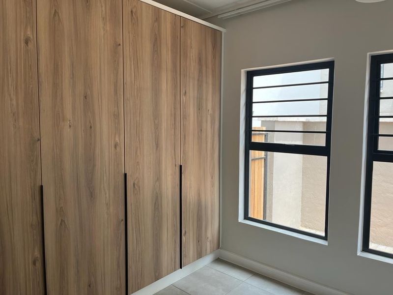 A bright bedroom featuring modern wood-finish built-in cupboards, tiled flooring, and large windows with black aluminium frames and security bars.